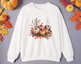 Könnte beinhalten: Ein weißes Sweatshirt mit einem Aquarelldesign von Kürbissen, Herbstblättern und einer dampfenden Tasse. Das Design umfasst orange, weiße und braune Kürbisse mit Akzenten aus grünen und roten Blättern und einer karierten Decke.