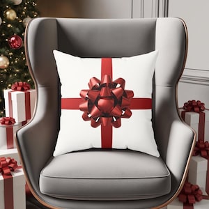 May include: A white square cushion featuring a large red bow and red ribbon cross design. The cushion is on a grey armchair, with wrapped gift boxes and a Christmas tree in the background.