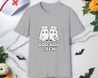 Pode incluir: Camiseta cinza claro com um desenho de dois fantasmas com chapéus de enfermeira e o texto "BOO BOO CREW". Decorações de Halloween, incluindo um chapéu de enfermeira, cercam a camiseta.