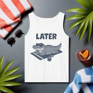 Könnte beinhalten: Weißes Tanktop mit dem Wort "LATER" in dunkelblauen Blockbuchstaben. Die Grafik zeigt ein graues Alligator mit Sonnenbrille, der sich auf einem Strandtuch mit einem Getränk entspannt. Sonnenbrille und Strandtuch liegen neben dem Tanktop.