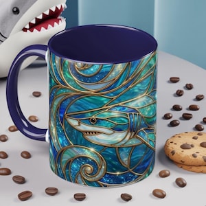 May include: A ceramic mug with a stained-glass design featuring a shark. The mug has a navy blue interior and handle, and the shark design is in shades of blue, teal, and gold. The mug is surrounded by coffee beans and a cookie.