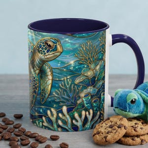 May include: A ceramic mug with a blue and white exterior and a navy blue interior. The mug features a detailed illustration of a sea turtle, coral, and ocean waves in shades of blue, teal, and gold. The mug is next to a plush turtle and chocolate chip cookies.