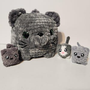 May include: A family of gray crocheted cat plushies. The large cat has green eyes and black whiskers. Three smaller cats are also present, one with a white and gray face, and two solid gray cats. All have embroidered facial features.