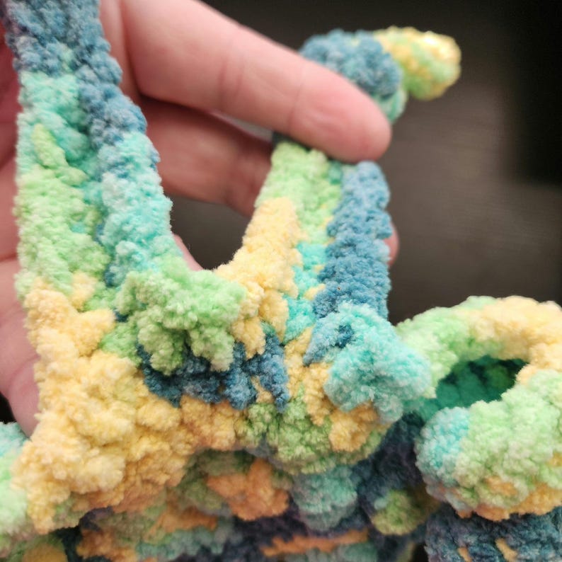 May include: Close-up of a handmade, colorful crochet item. The yarn is a soft, textured material in shades of blue, green, and yellow. The item appears to be a decorative piece or part of a larger craft project.