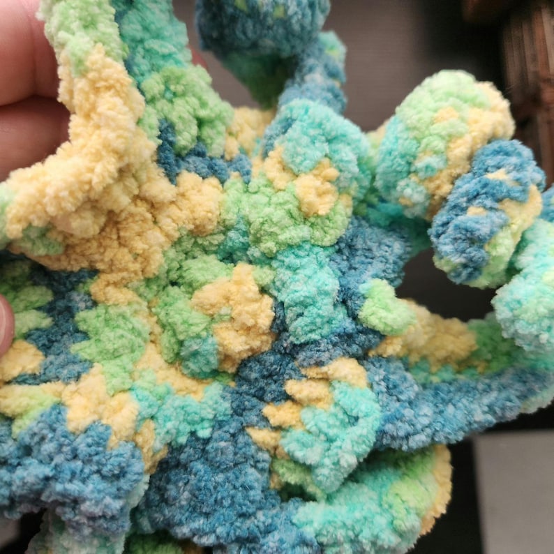 May include: Close-up of a handmade, textured crochet item in shades of blue, green, and yellow. The soft, plush yarn is intricately looped, creating a unique, organic shape. The color palette evokes a natural, coastal theme.