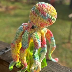 May include: A colorful, crocheted octopus toy with a vibrant mix of yellow, pink, blue, and green yarn. The plush octopus has eight curly tentacles and a round head. The toy is sitting on a wooden surface with a green object behind it.