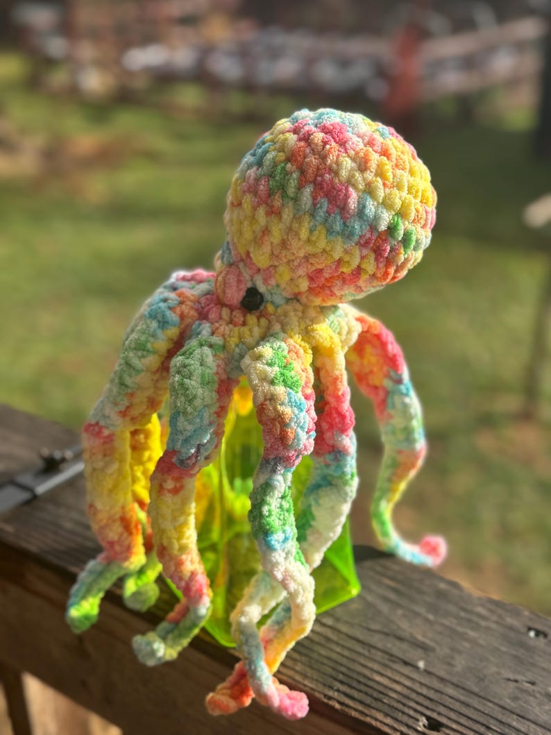 May include: A colorful, crocheted octopus toy with a vibrant mix of yellow, pink, blue, and green yarn. The plush octopus has eight curly tentacles and a round head. The toy is sitting on a wooden surface with a green object behind it.