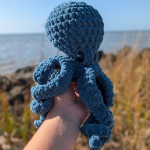 May include: A handmade, blue crochet octopus with a textured surface. The plush toy has a round head, eight curled tentacles, and a pair of black button eyes. The octopus is held up against a backdrop of the ocean and a clear blue sky.