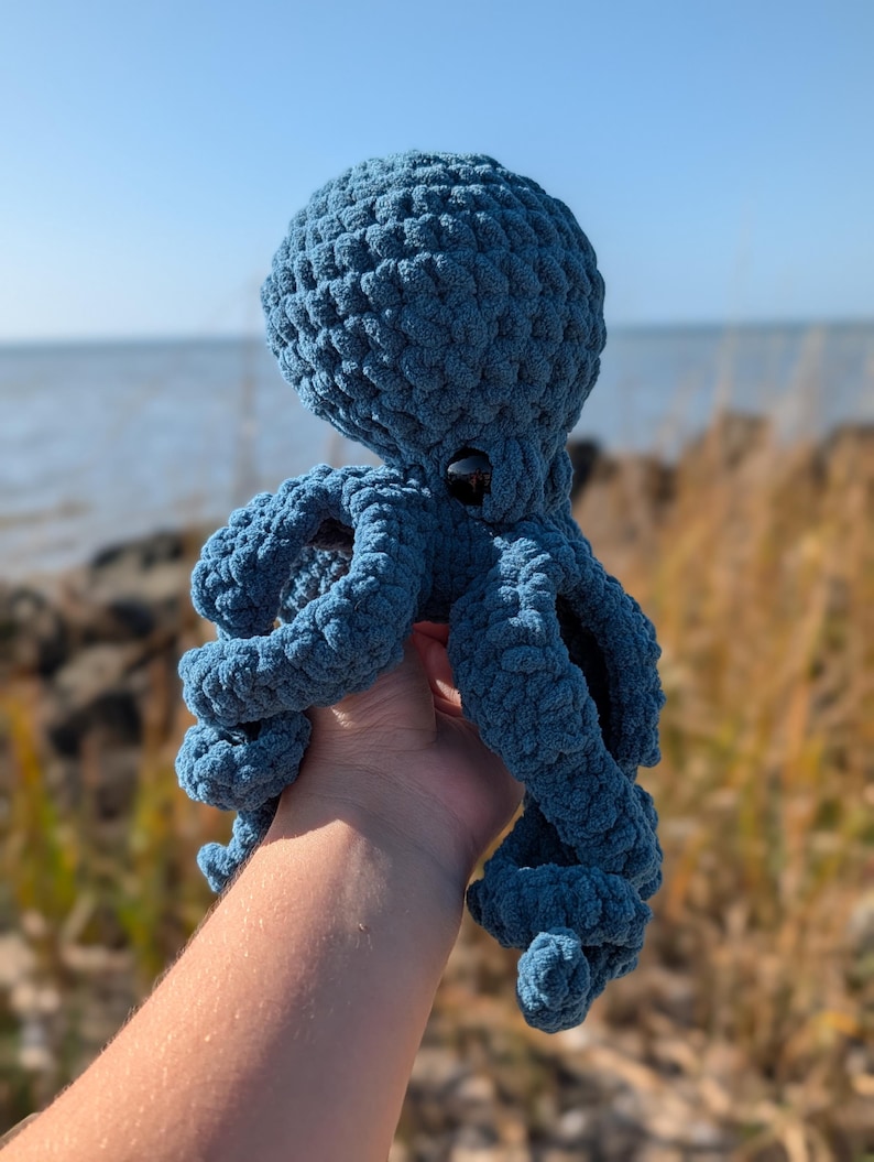 May include: A handmade, blue crochet octopus with a textured surface. The plush toy has a round head, eight curled tentacles, and a pair of black button eyes. The octopus is held up against a backdrop of the ocean and a clear blue sky.
