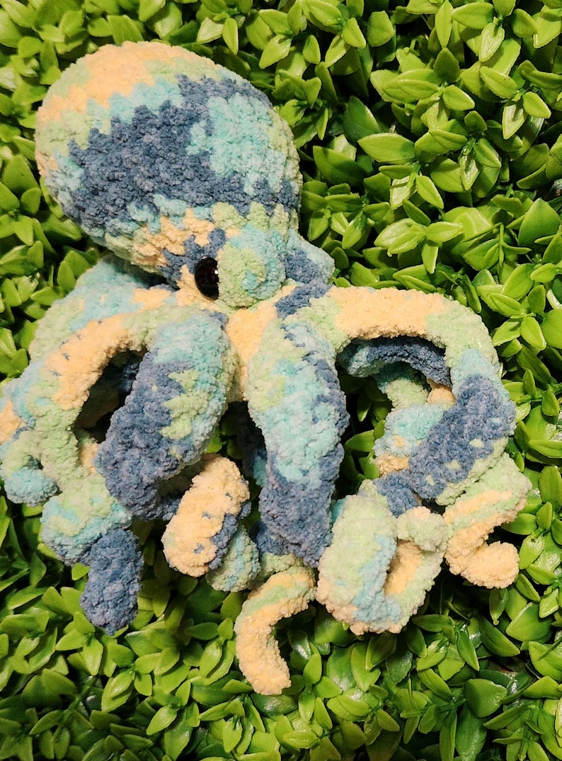 May include: A handmade plush octopus in shades of blue, green, and yellow. The soft, textured toy has eight curled tentacles and a round head with black eyes. The octopus is set against a background of green foliage.