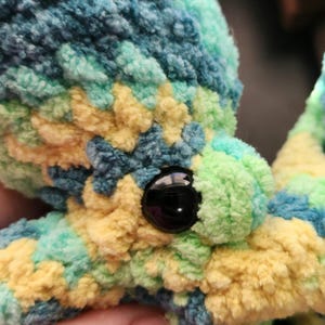May include: A close-up of a handmade, plush octopus toy. The octopus is crafted from soft, textured yarn in a vibrant mix of colors, including yellow, teal, blue, and green. It has a large, black, button-like eye.