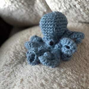 May include: A small, handmade blue crochet octopus with eight tentacles and a round head. The octopus has a black button eye and is resting on a soft, white, textured surface. The crochet work is detailed and the color is a muted blue.
