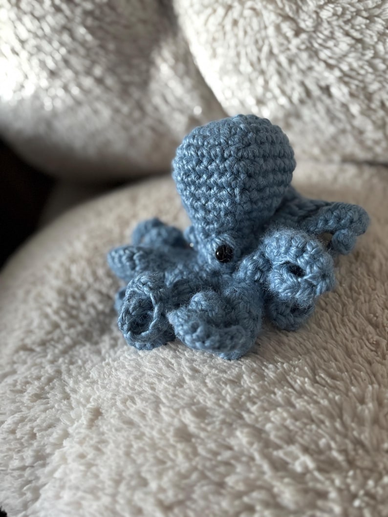 May include: A small, handmade blue crochet octopus with eight tentacles and a round head. The octopus has a black button eye and is resting on a soft, white, textured surface. The crochet work is detailed and the color is a muted blue.