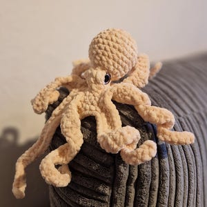 May include: A plush, peach-colored octopus toy with eight long, curly tentacles. The stuffed animal has a round head and small, black eyes. The octopus is made of a soft, textured material and is resting on a gray, corduroy surface.
