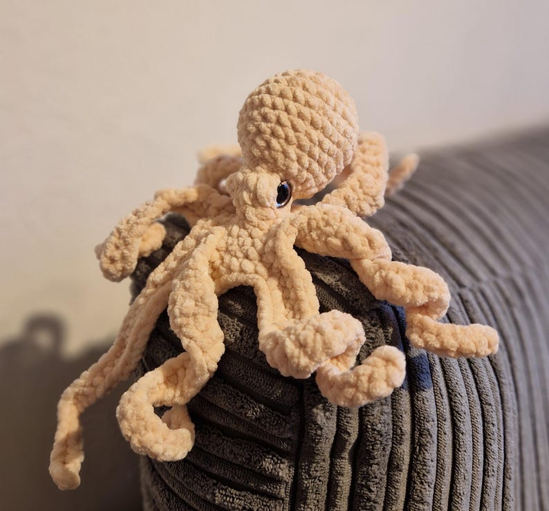 May include: A plush, peach-colored octopus toy with eight long, curly tentacles. The stuffed animal has a round head and small, black eyes. The octopus is made of a soft, textured material and is resting on a gray, corduroy surface.