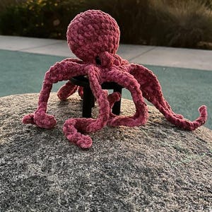 May include: A handmade, plush, pink octopus toy with eight long tentacles. The octopus is sitting on a gray rock, with a blurred background of trees and a sunset.