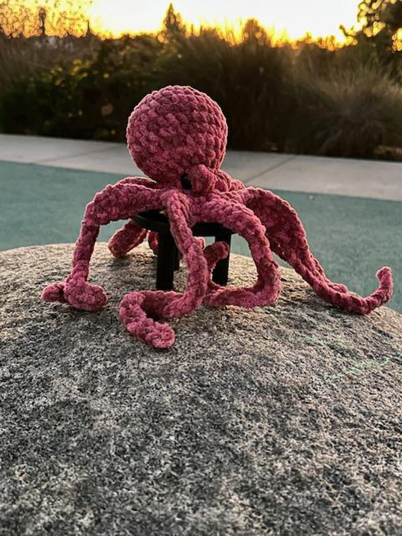 May include: A handmade, plush, pink octopus toy with eight long tentacles. The octopus is sitting on a gray rock, with a blurred background of trees and a sunset.
