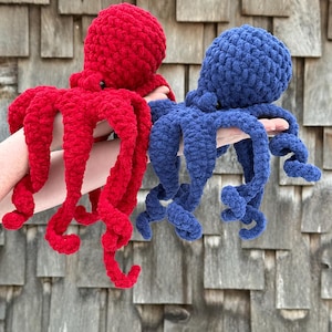 May include: Two crocheted octopus plush toys, one red and one blue, are held up against a weathered wood background. The plushies have rounded heads and long, curly tentacles. The red octopus is on the left, and the blue one is on the right.