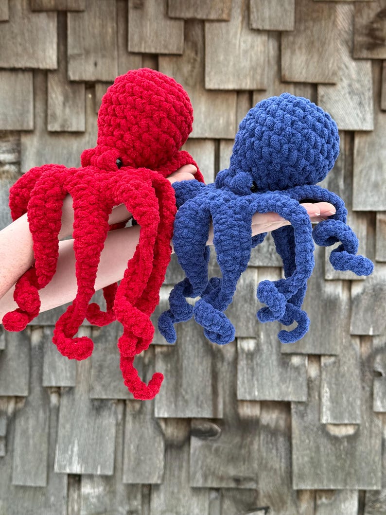 May include: Two crocheted octopus plush toys, one red and one blue, are held up against a weathered wood background. The plushies have rounded heads and long, curly tentacles. The red octopus is on the left, and the blue one is on the right.