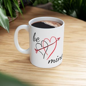 May include: White ceramic coffee mug with a black handle. The mug has a black and red design with two hearts and an arrow, and the text "be mine".