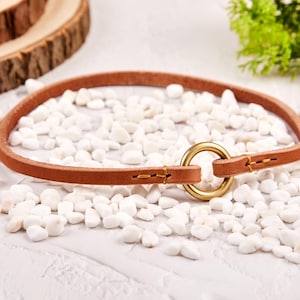 Peut inclure: Un collier de chien en cuir marron avec un anneau en laiton. Le collier est posé sur une surface blanche avec des pierres blanches.