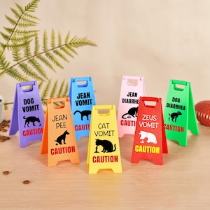 May include: A collection of colorful, miniature caution signs with humorous pet-related warnings. Each sign features a different color and text, including "Dog Vomit," "Cat Vomit," and "Dog Diarrhea." A football and pet food bowl are also in the image.