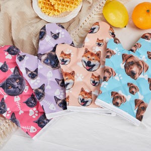 May include: Four oven mitts with different pet faces printed on them. The mitts are pink, light purple, light blue, and peach. Each mitt has a white border.
