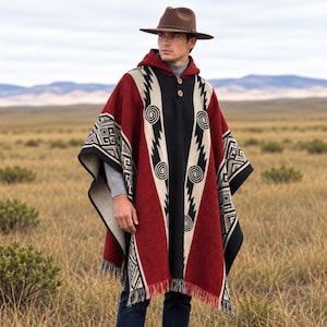 May include: A hooded poncho in deep red, black, and cream featuring geometric patterns and spiral motifs. The garment includes a two-button closure at the neckline and a fringed hem. It is worn with a brown fedora hat, a grey sweater, and blue jeans.