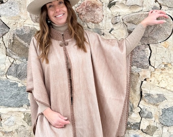 May include: A beige poncho featuring a high collar with a textured knit trim, a single toggle button closure at the neck, and a decorative stitched border along the edges. The garment is worn over a matching beige turtleneck sweater and blue jeans.