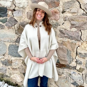 Op de afbeelding: Een crèmekleurige poncho met een decoratieve rand en twee houten knopen. De poncho wordt gedragen over een beige coltrui en een blauwe spijkerbroek. Een beige hoed wordt gedragen. De achtergrond is een stenen muur.