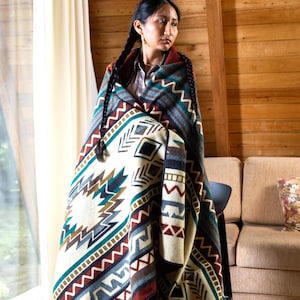 May include: A woven blanket with a geometric pattern in cream, teal, brown, and red. The blanket features a central design with diamond and arrow shapes, and zig-zag borders. The blanket is draped over a person, showcasing its size and design.