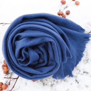 May include: A rolled navy blue scarf with a fringed edge, presented against a white backdrop. Sprigs of berries are arranged around the scarf. The deep blue colour and soft texture of the scarf are highlighted, suggesting a cosy accessory.