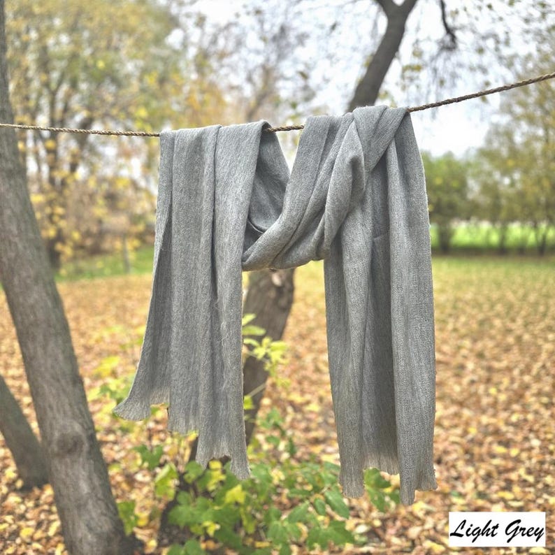 May include: A light grey scarf hangs from a washing line, with the text "Light Grey" in the bottom right corner. The scarf is made of a soft material and has a fringed edge. The background is a blurred outdoor scene.