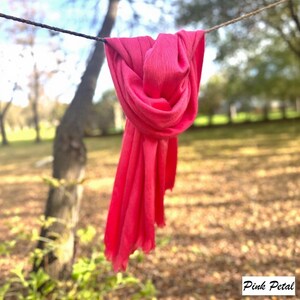 May include: A bright pink scarf hangs from a washing line, with the text "Pink Petal" in the lower right corner. The scarf's fabric appears soft and flowing. The background shows a blurred park scene with trees and grass.