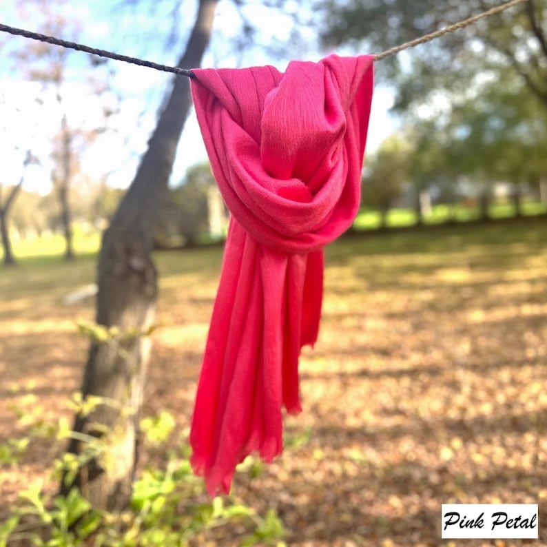 May include: A bright pink scarf hangs from a washing line, with the text "Pink Petal" in the lower right corner. The scarf's fabric appears soft and flowing. The background shows a blurred park scene with trees and grass.
