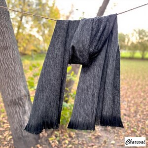 May include: A dark grey scarf hangs from a washing line. The scarf has a textured finish and a fringed edge. The word "Charcoal" is written in a script font in the bottom right corner. The background is blurred, with trees and foliage.