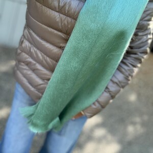 May include: A light green scarf with a fringed edge is held against a brown quilted vest and blue jeans. The scarf's color transitions from a darker green to a lighter, brighter shade. The overall look suggests a casual, everyday style.