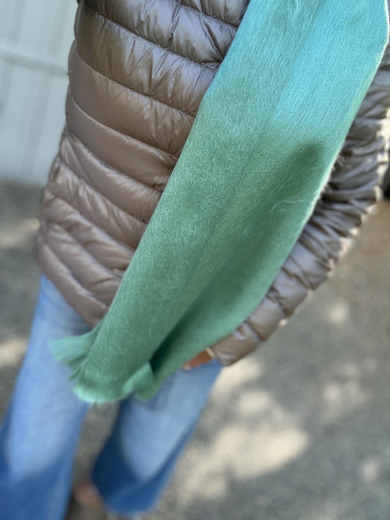 May include: A light green scarf with a fringed edge is held against a brown quilted vest and blue jeans. The scarf's color transitions from a darker green to a lighter, brighter shade. The overall look suggests a casual, everyday style.