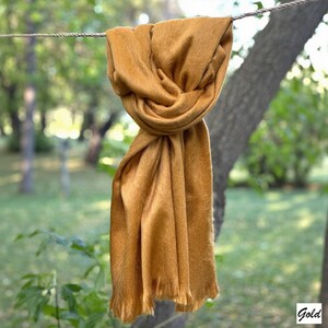 May include: A mustard-coloured scarf is draped over a rope, with a blurred background of trees and greenery. The scarf has a soft, textured appearance and fringed edges. The word "Gold" is in the bottom right corner.