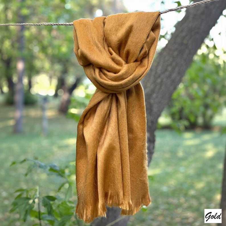 May include: A mustard-coloured scarf is draped over a rope, with a blurred background of trees and greenery. The scarf has a soft, textured appearance and fringed edges. The word "Gold" is in the bottom right corner.