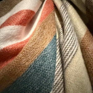 May include: A soft, woven textile featuring a pattern of wide, diagonal stripes in shades of terracotta orange, muted teal, tan, and cream. The fabric has a visible, slightly fuzzy texture with sections displaying a fine, dark brown pinstripe detail.