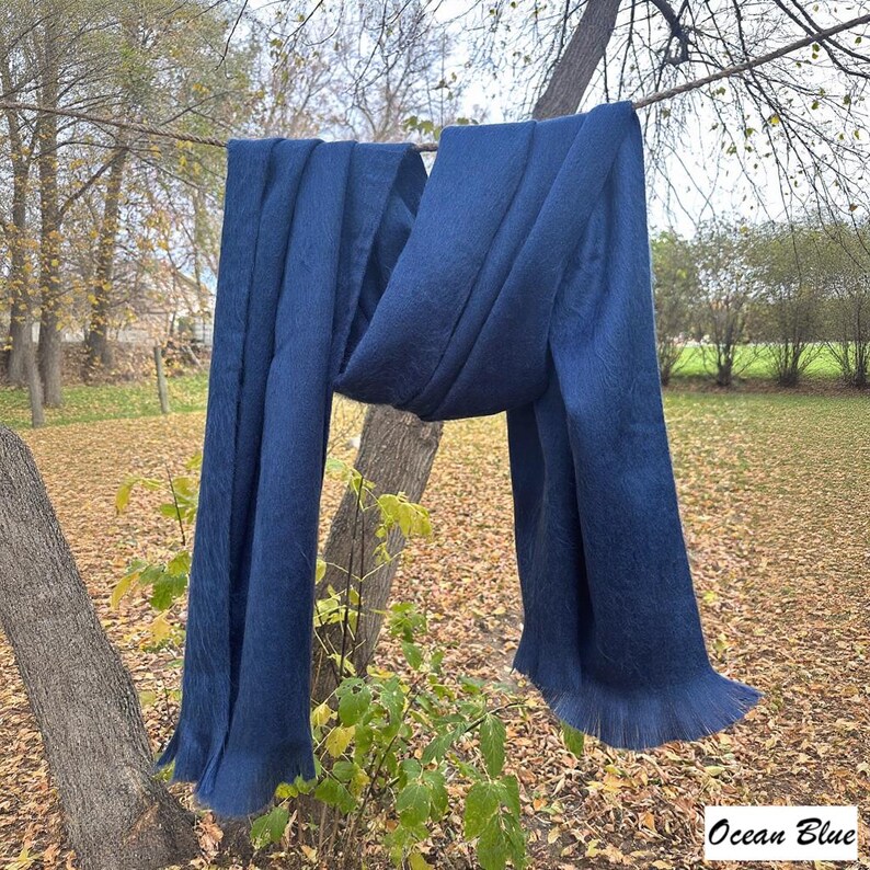 May include: A long, navy blue scarf is draped over a tree branch. The scarf has a fringed edge and is labelled "Ocean Blue" in the bottom right corner. The scarf is made of a soft material and is hanging outside in a natural setting.