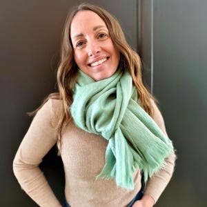May include: A woman wearing a light green scarf draped around her neck. The scarf has a fringed edge and is paired with a tan ribbed sweater. The woman is smiling, with long brown hair.