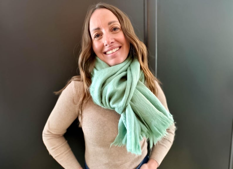 May include: A woman wearing a light green scarf draped around her neck. The scarf has a fringed edge and is paired with a tan ribbed sweater. The woman is smiling, with long brown hair.