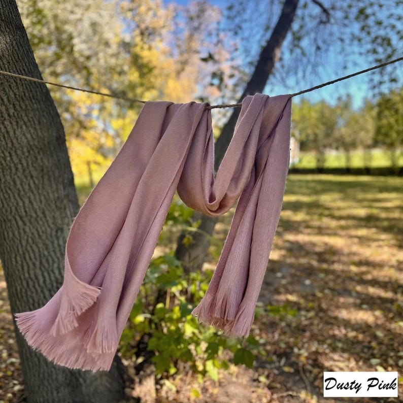 May include: A dusky pink scarf with fringed edges hangs from a line outdoors. The scarf is draped in soft folds, showing its texture and colour. The text "Dusty Pink" is in the bottom right corner.