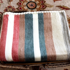 May include: A rectangular throw blanket with vertical stripes in red, white, brown, teal, and grey. The blanket is folded and placed inside a woven basket. The blanket's edges are finished with a white trim.