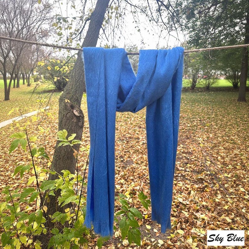 May include: A sky blue scarf hangs from a rope, set against a backdrop of trees and fallen leaves. The scarf is a vibrant blue and appears to be made of a soft fabric. The text "Sky Blue" is visible in the bottom right corner.