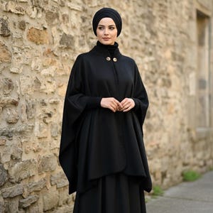 May include: A black cape-style coat with gold-colored buttons, worn over a black turtleneck and a matching black head covering. The outfit is paired with a long black skirt. The background is a stone wall.