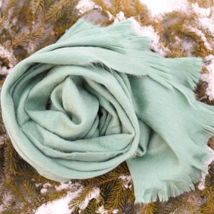 May include: A soft, mint green scarf is artfully arranged on a bed of evergreen branches dusted with snow. The scarf has a fringed edge and is made of a fuzzy material, perfect for cold weather. The scarf is a solid color.