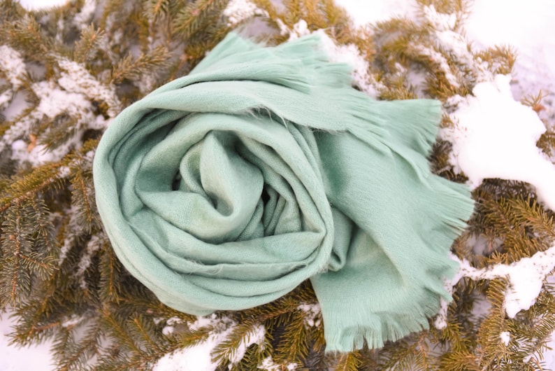 May include: A soft, mint green scarf is artfully arranged on a bed of evergreen branches dusted with snow. The scarf has a fringed edge and is made of a fuzzy material, perfect for cold weather. The scarf is a solid color.